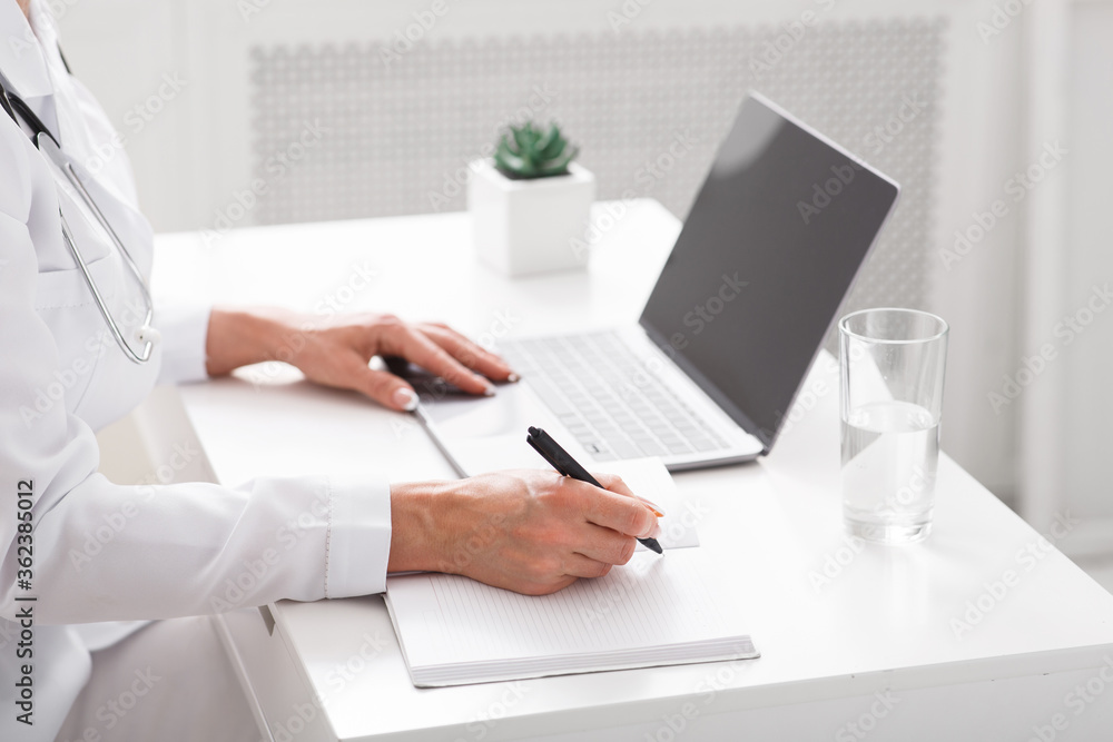 Online communication with patient and notes. Female doctor writes in notebook at table with laptop Online communication with patient and notes. Female doctor writes in notebook at table with laptop