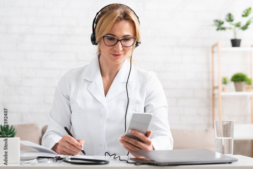 Writes patients to visit. Woman in white coat with headphones, smartphone and laptop makes notes in notebook Writes patients to visit. Woman in white coat with headphones, smartphone and laptop makes notes in notebook