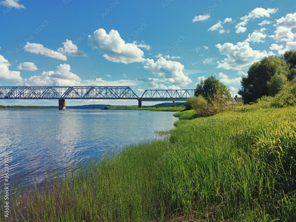 Naklejka premium green river bank in front of a bridge against a blue sky with clouds on a sunny day