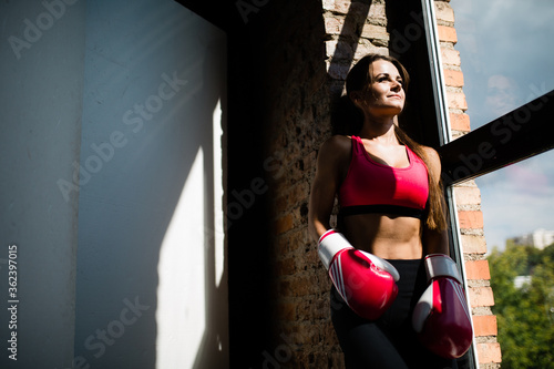 Portrait of a boxer girl in boxing gloves who stands next to a panoramic window under the morning sun