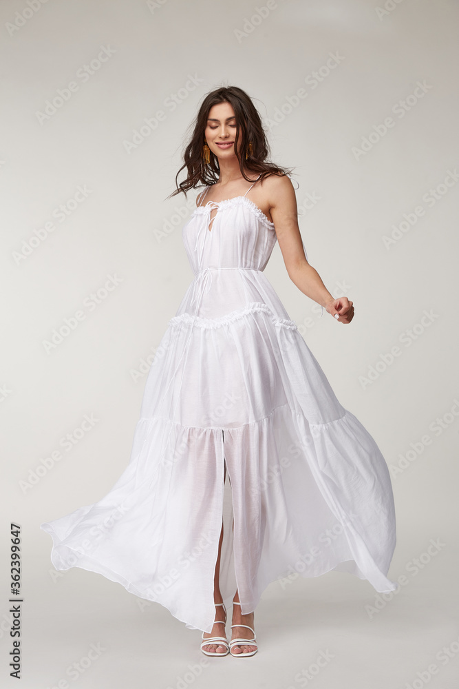 Fototapeta premium Beautiful woman fashion model makeup brunette hair perfect body shape tanned skin wear clothes summer collection organic long white silk dress stylish sandals shoes, accessory jewelry earrings date.