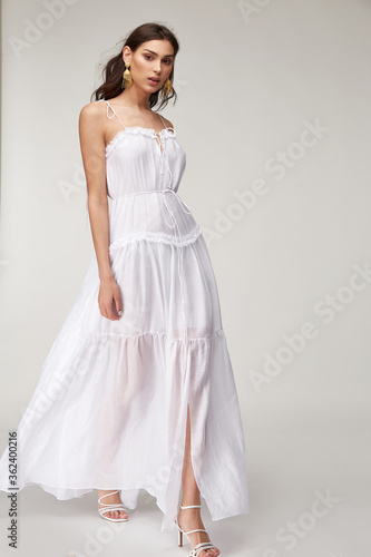 Fotografie Beautiful woman fashion model makeup brunette hair perfect body shape tanned skin wear clothes summer collection organic long white silk dress stylish sandals shoes, accessory jewelry earrings date