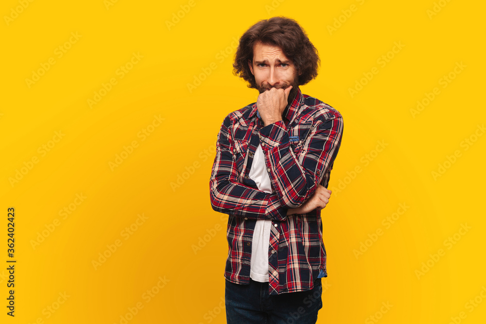 A young man with long curly hair and a beard is worried, worried, bites his fist, yellow background. A young man with long curly hair and a beard is worried, worried, bites his fist, yellow background.