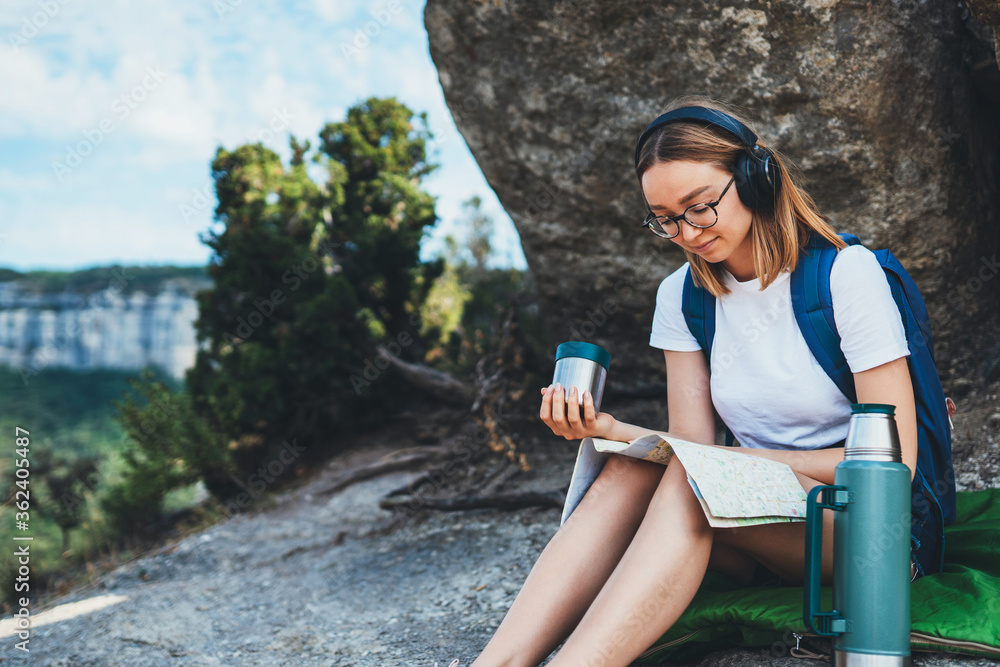 hiker girl with backpack and сup of drink looks at map mountains plans trip route, female person in glasses planning hiking while listening to music in wireless headphones and relaxing during journey