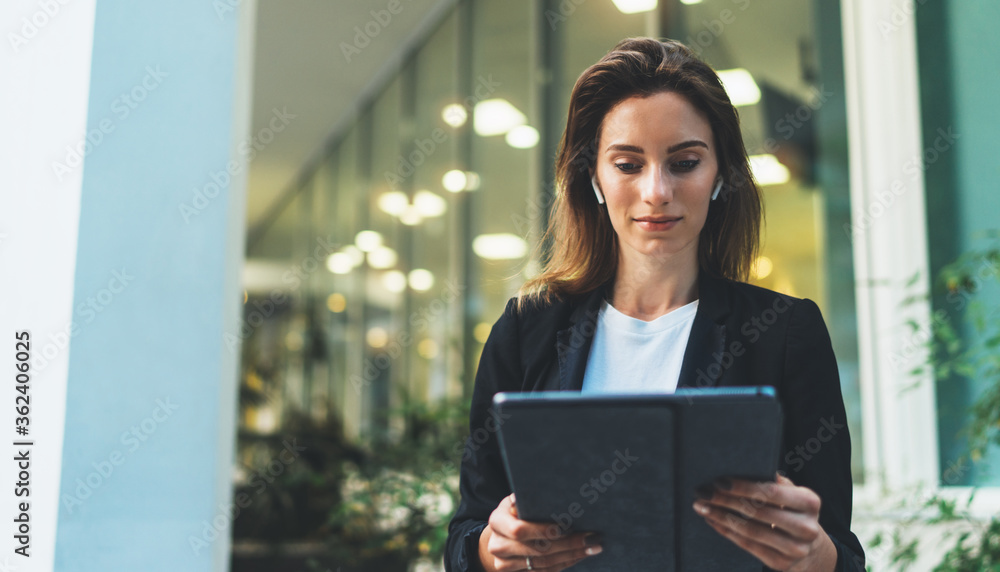 Successful female banker using tablet and wireless earphones outdoors near his office background lights, portrait young woman professional manager working on touch pad in evening city Successful female banker using tablet and wireless earphones outdoors near his office background lights, portrait young woman professional manager working on touch pad in evening city
