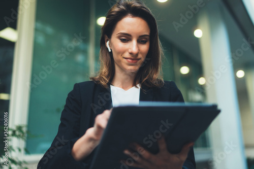 Successful female banker using tablet and wireless earphones outdoors near his office background lights, portrait young woman professional manager working on touch pad near skyscraper in evening city photo