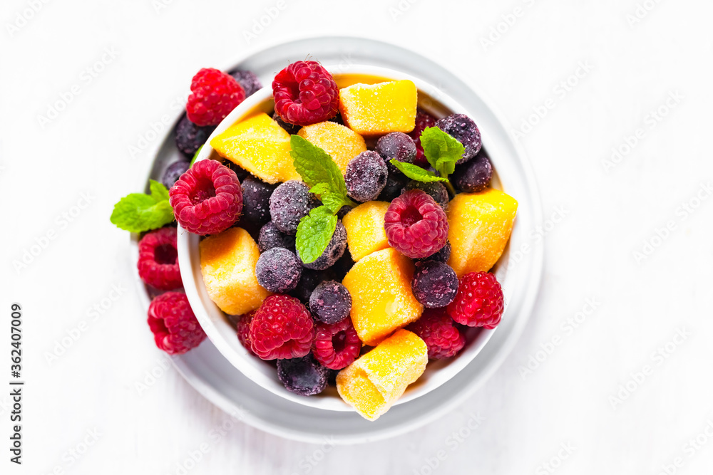 Frozen fruits: mango, raspberry, blueberry with fresh green mint, white bowl. Concept of healthy eating, low calories dessert, summer refreshing meal. Light background copy space. Flat lay top view