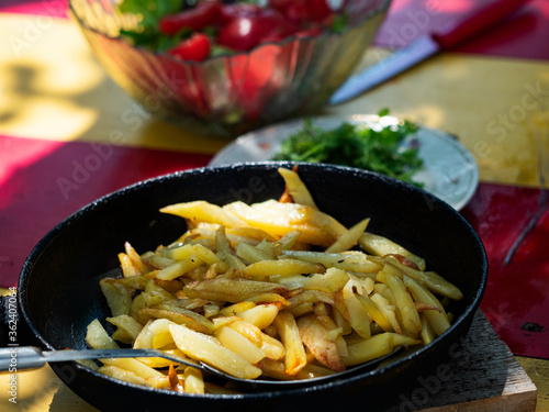 Fried potatoes in a pan on the table on the background of a bowl with salad