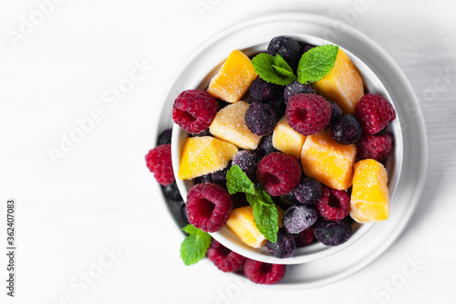 Frozen fruits  mango  raspberry  blueberry with fresh green mint  white bowl. Concept of healthy eating  low calories dessert  summer refreshing meal. Light background copy space. Flat lay top view