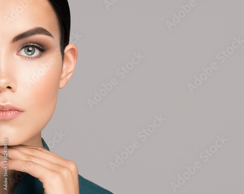 Close up crop view of young brunette Caucasian woman with fresh skin looking in front of the camera with a hand on a chin. Isolated on grey background with copy space for text. Cosmetics business look © Helena Lansky