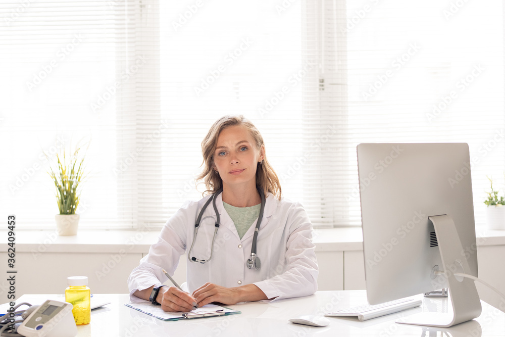 Pretty young successful female clinician in whitecoat making notes by desk Pretty young successful female clinician in whitecoat making notes by desk