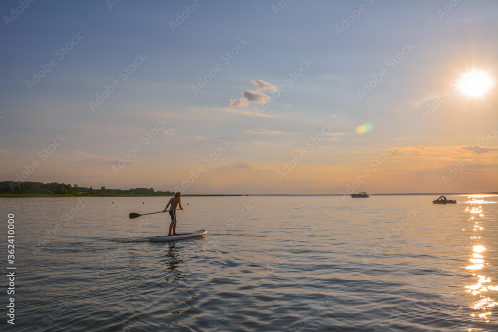 Naklejka premium Young boy on the lake is rowing on the inflatable sup towards the sunset
