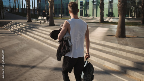 Back view of guy with backpack walking along street after training outdoor photo