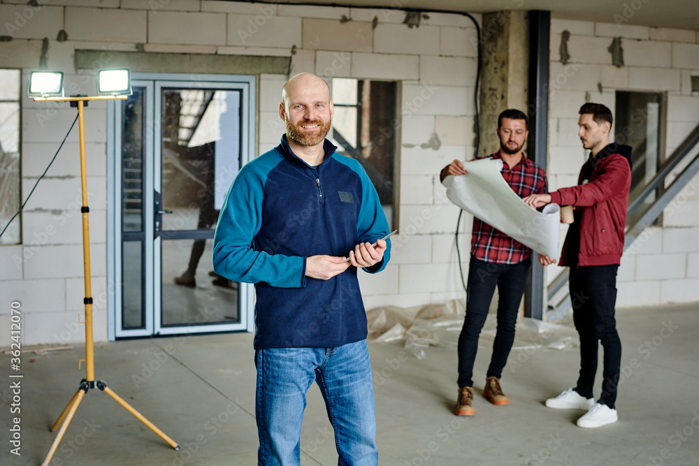 Fototapeta premium Young bald architect or contractor looking at you on background of colleagues