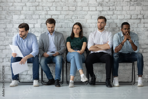 Five multi racial young people applicants sitting in line on chairs in office corridor feels nervous unsure job interview success, highly competitive job market, tension rises due to waiting concept