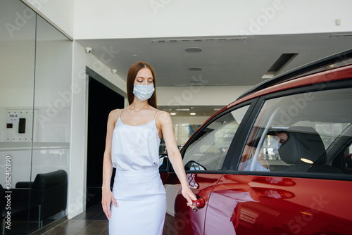 A young pretty girl inspects a new car at a car dealership in a mask during the pandemic. The sale and purchase of cars, in the period of pandemia