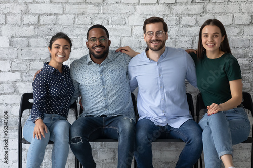 Four multi ethnic students friendly office employees sit together on chairs embracing smiling looking at camera. Warm relations between company members, amity and friendship, racial equality concept