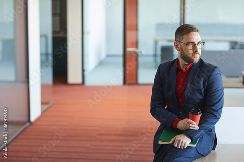 High angle portrait of successful bearded businessman looking away while sitting on desk with cup of coffee in modern office interior, copy space