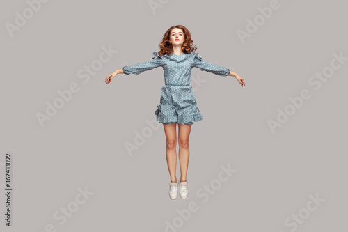 Full length happy calm pretty girl in vintage ruffle dress levitating hovering in mid-air with raised hands as wings, jumping trampoline or flying up. indoor studio shot isolated on gray background photo