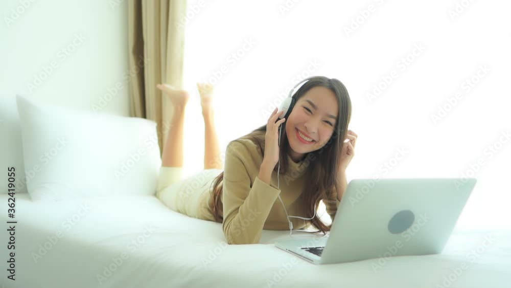 Happy, smiling young Asian woman laying on bed near window with bright light bounces and dances to music streaming on laptop computer to wireless headphones. Happy, smiling young Asian woman laying on bed near window with bright light bounces and dances to music streaming on laptop computer to wireless headphones.