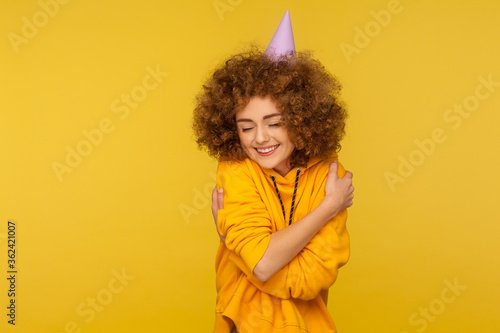 Portrait of selfish curly-haired hipster girl with funny cone hat embracing herself and smiling with pleasure expression, self-loving and complacency concept. studio shot isolated on yellow background
