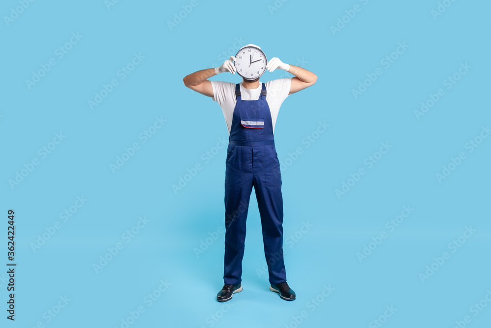 Naklejka premium Full length anonymous labourer in uniform covering face with clock. Advertisement of professional house repair services on time, punctual courier delivery. studio shot isolated on blue background