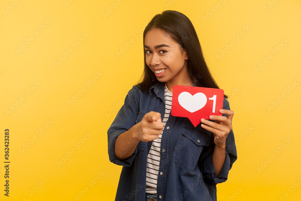 Hey you! Portrait of playful girl in denim shirt pointing to camera and showing heart Like icon, follower notification symbol, click button for social media. studio shot isolated on yellow background