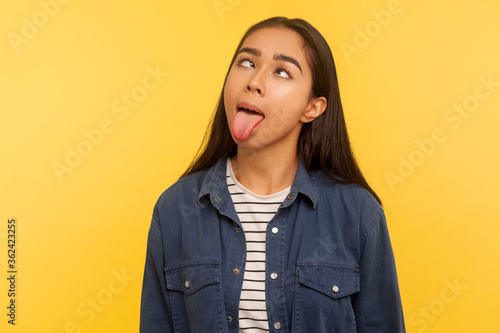 Konstfotografi Portrait of funny dumb crazy girl in denim shirt showing tongue out and looking cross-eyed with silly brainless expression, aping fooling around