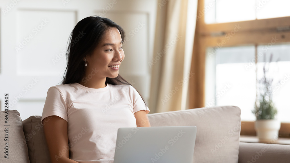 Smiling beautiful young asian ethnicity woman sitting on sofa with computer on lap, thinking of daily work, planning freelance working schedule, dreaming of vacation or visualizing alone at home. Smiling beautiful young asian ethnicity woman sitting on sofa with computer on lap, thinking of daily work, planning freelance working schedule, dreaming of vacation or visualizing alone at home.