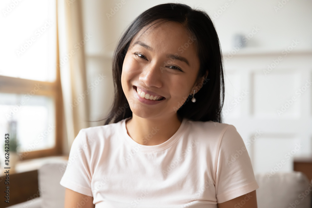 Head shot close up beautiful smiling vietnamese ethnicity young woman sitting on sofa, looking at camera. Happy biracial millennial lady blogger recording video or holding call remotely from home. Head shot close up beautiful smiling vietnamese ethnicity young woman sitting on sofa, looking at camera. Happy biracial millennial lady blogger recording video or holding call remotely from home.