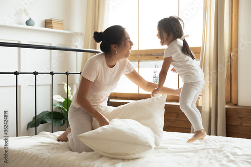 Excited vietnamese ethnicity female family in pajamas having fun on comfortable bed, playing pillow fight, enjoying energetic morning time after waking up together, laughing joking in bedroom.