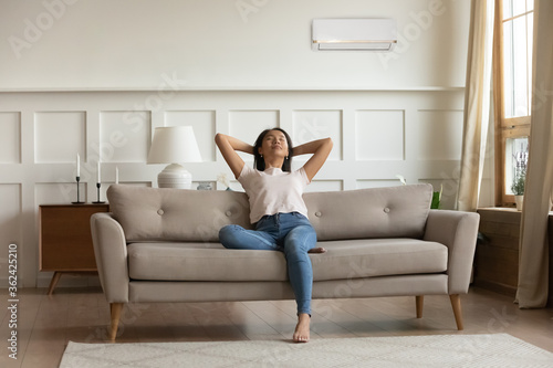 Happy young calm woman relaxing on sofa, breathing fresh air, satisfied with comfortable indoors temperature. enjoying spending hot summer time in cooled living room, front full length view.