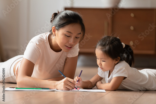 Affectionate caring vietnamese ethnicity woman lying on floor with cute small preschool biracial child daughter, involved in hand drawing pictures together in paper album, childcare hobby pastime.