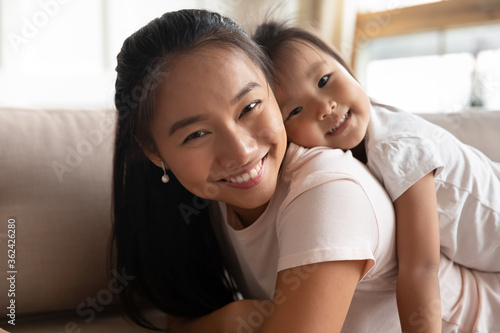 Head shot portrait smiling attractive young asian mother resting on sofa, holding on back cute small preschool vietnamese ethnicity child daughter, enjoying relaxed free time, looking at camera.