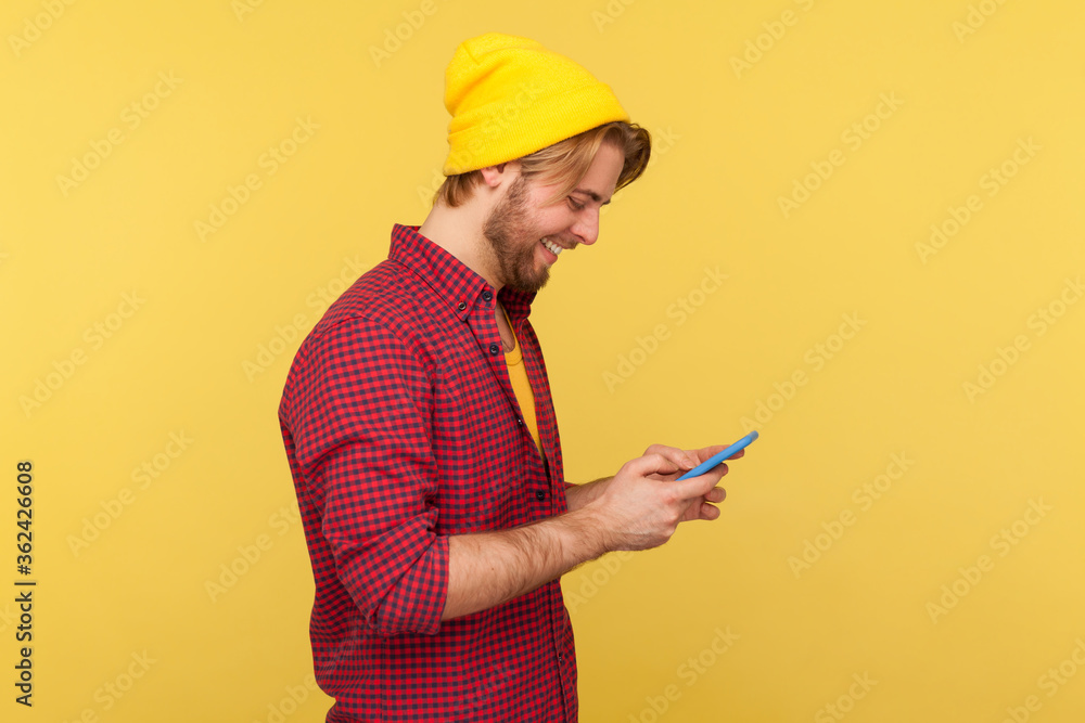 Side view happy trendy guy in hipster beanie hat and shirt using cell phone, texting or reading message, smiling while chatting in social network, enjoying mobile app. indoor studio shot isolated Side view happy trendy guy in hipster beanie hat and shirt using cell phone, texting or reading message, smiling while chatting in social network, enjoying mobile app. indoor studio shot isolated