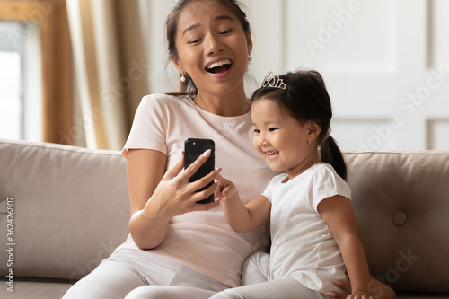Happy young asian mother showing funny educational application on mobile phone to interested cute small kid daughter, relaxing together on sofa, excited family using digital gadget, watching videos.