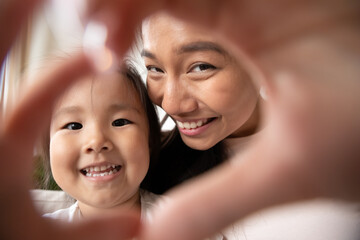 Close up head shot charming little preschool asian baby girl making heart gesture with smiling beautiful vietnamese ethnic mother, showing love, care and devotion, happy family web camera view.