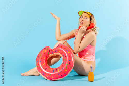 Full length, indignant angry woman in bathing suit sitting with rubber ring, cocktail and talking on phone handset with resentful annoyed expression. indoor studio shot isolated on blue background © khosrork
