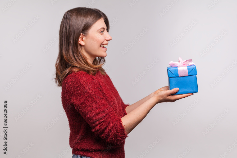 Please, take this christmas present! Side view, happy girl in shaggy sweater giving decorated gift box and greeting, congratulating on new year, x-mas holidays. studio shot isolated on gray background Please, take this christmas present! Side view, happy girl in shaggy sweater giving decorated gift box and greeting, congratulating on new year, x-mas holidays. studio shot isolated on gray background