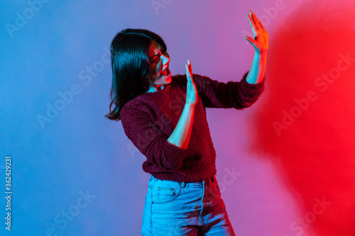 I am afraid! Side view, portrait of scared woman standing with raised hands, hiding from fear and screaming in horror, looking frightened as notices something terrible. colorful neon light studio shot
