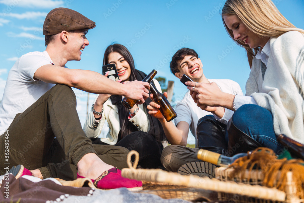 Happy multiracial friends having fun on a picnic - Youth toasting with beer and using their smart phones - Friendship concept in the fresh entertainment mood - Focus on the youth ahead