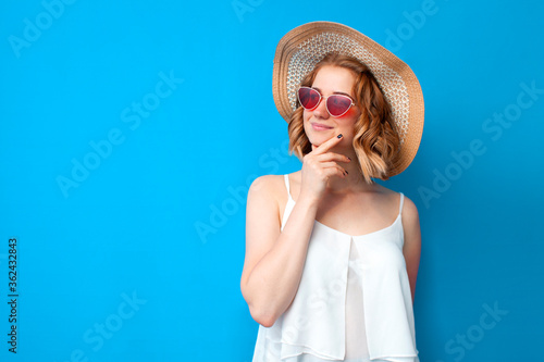 thoughtful girl in a sun hat and glasses thinks on a blue isolated background, woman in summer clothes, summer concept