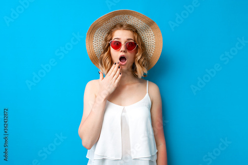 summer concept. shocked girl in a sun hat and glasses is surprised on a blue isolated background, emotional woman in summer clothes on vacation