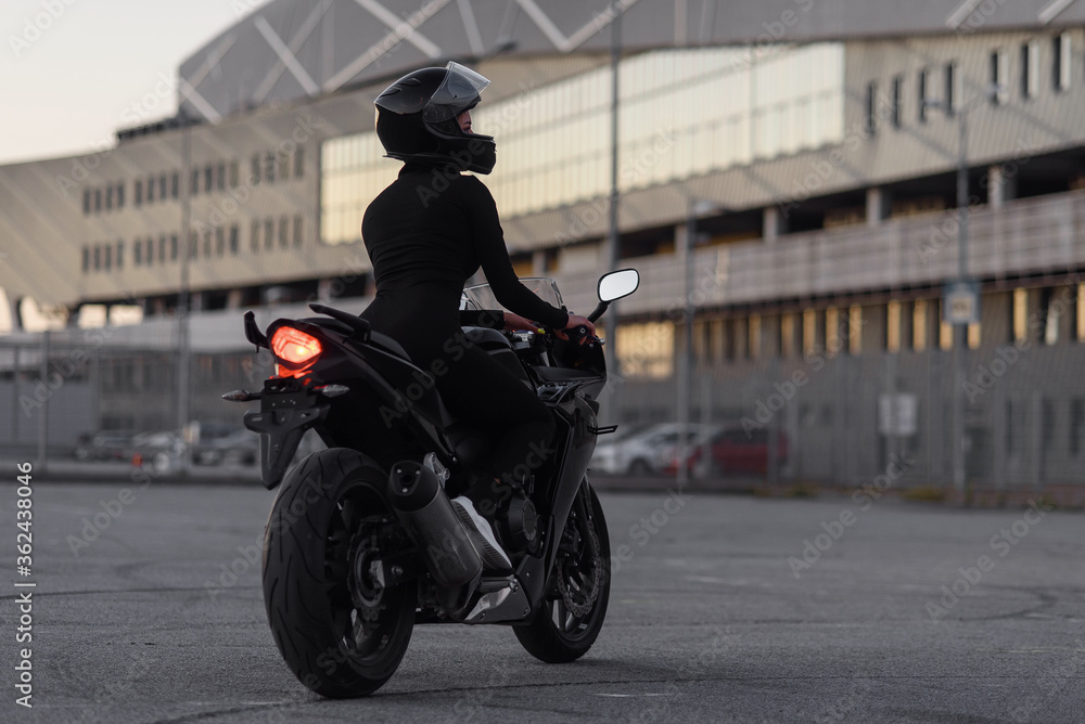 Back view of attractive young woman in black tight-fitting suit and full-face protective helmet rides on sports motorcycle at urban outdoors parking in the evening.