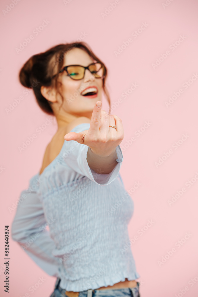 Cute eccentric provocative girl looking at camera, inviting with a finger. Over pink background. She wears glasses, two buns on her head and a shirt with an open top. Focus on a hand. Cute eccentric provocative girl looking at camera, inviting with a finger. Over pink background. She wears glasses, two buns on her head and a shirt with an open top. Focus on a hand.