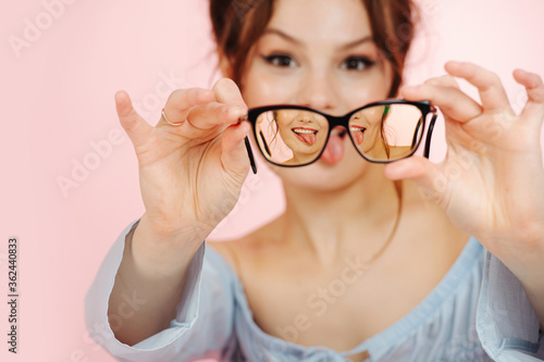 Eccentric young woman holding glasses in front of her face  sticking tongue out. Image triples in lenses. Over pink background.