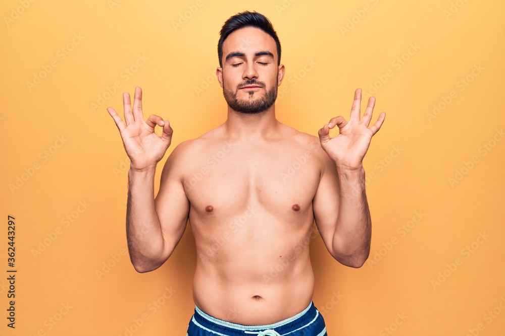 Young handsome man with beard wearing sleeveless t-shirt standing over yellow background relax and smiling with eyes closed doing meditation gesture with fingers. Yoga concept.