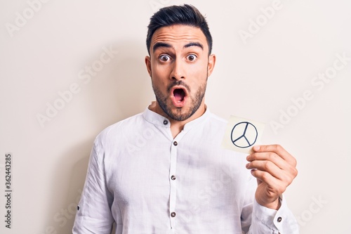 Young handsome man with beard holding reminder with peace symbol over white background scared and amazed with open mouth for surprise, disbelief face