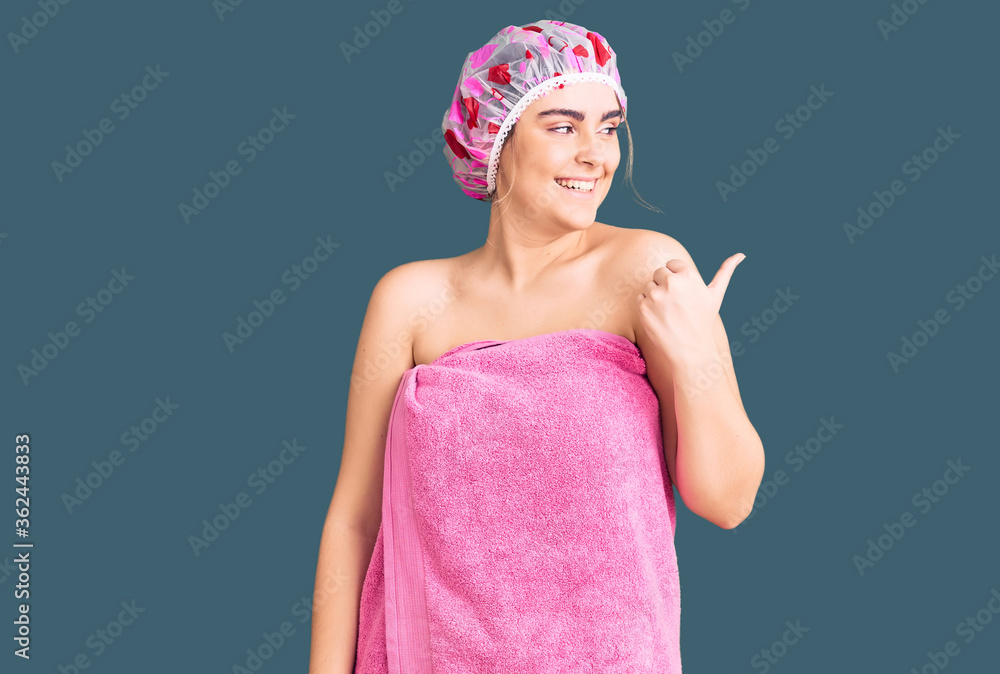 Young caucasian woman wearing shower cap and towel smiling with happy face looking and pointing to the side with thumb up. Young caucasian woman wearing shower cap and towel smiling with happy face looking and pointing to the side with thumb up.