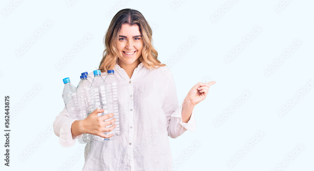 Obraz premium Young caucasian woman holding recycling plastic bottles smiling happy pointing with hand and finger to the side